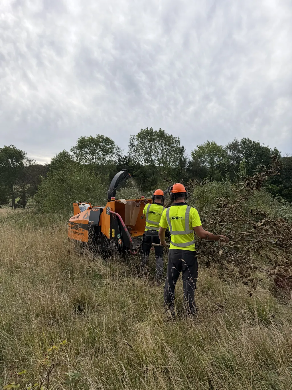 JRW Garden Services operatives in hi-vis and safety helmets feeding cut vegetation into a woodchipper during a site clearance